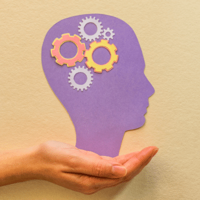 A hand holds a purple silhouette of a head with yellow and white gears inside, symbolizing thinking or mental processes.