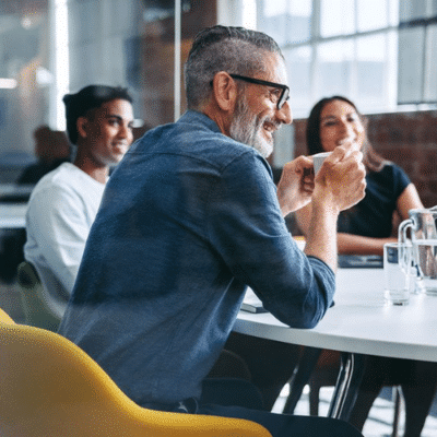 A white haired man with a beard drinks a coffee in a meeting with colleagues